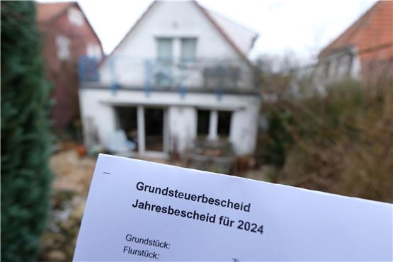 Ein Grundsteuerbescheid für 2024 wird vor einem Privatgrundstück hochgehalten. Foto: Bernd Weißbrod/dpa